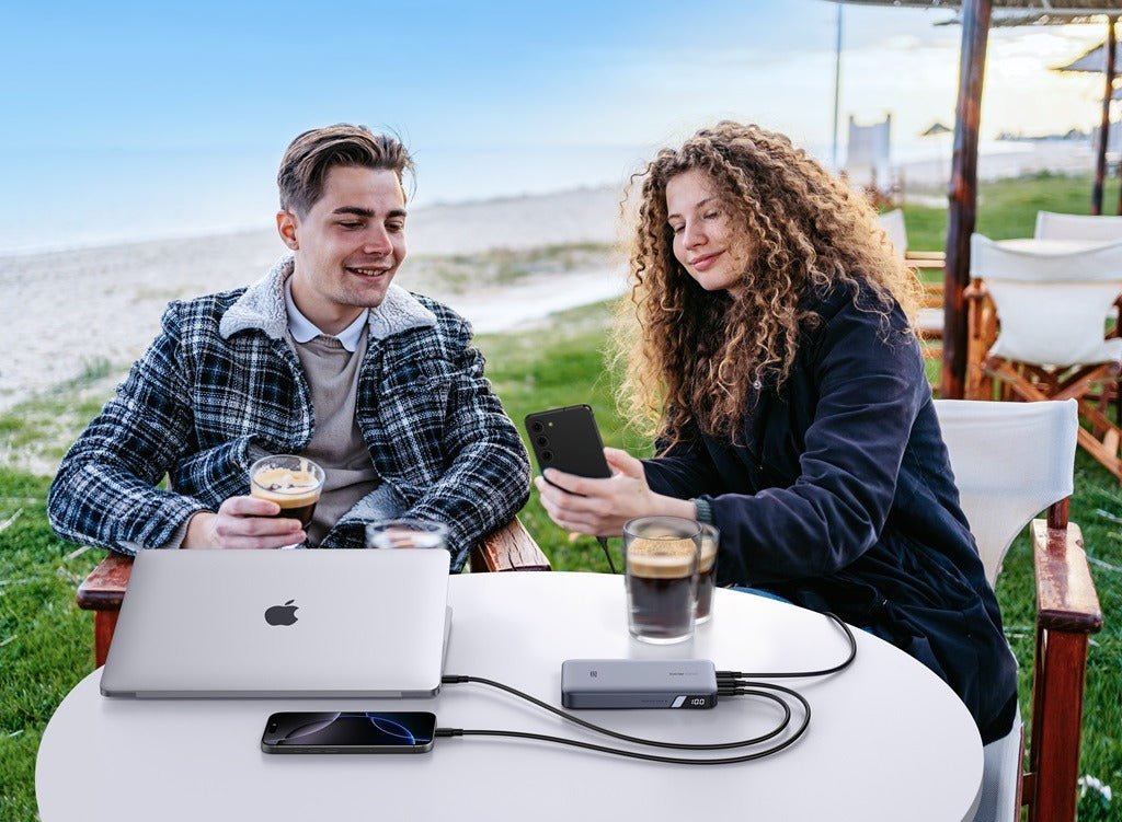 Two people outdoors using a power bank to charge a laptop and smartphones at a table