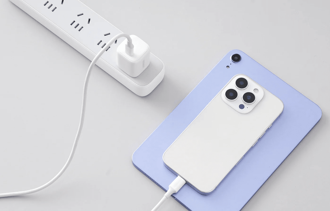 White USB-C charger plugged into a power strip, charging a white smartphone resting on a light blue tablet.