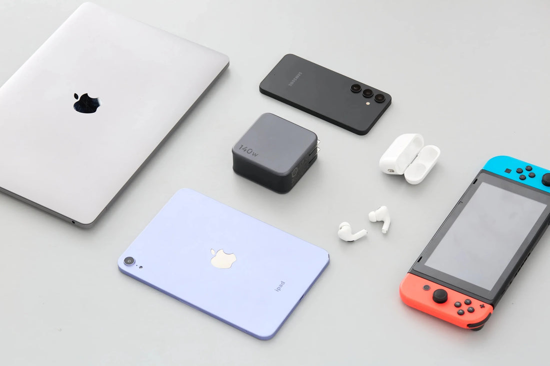 Flat lay of electronic devices including a laptop, smartphone, tablet, charger, wireless earbuds, and handheld gaming console on a gray surface