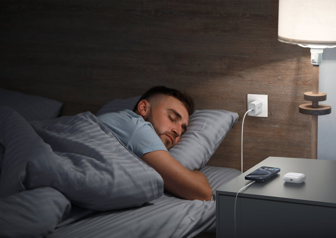 A man sleeping in bed at night while his smartphone charges on a bedside table next to wireless earbuds and a wall charger.